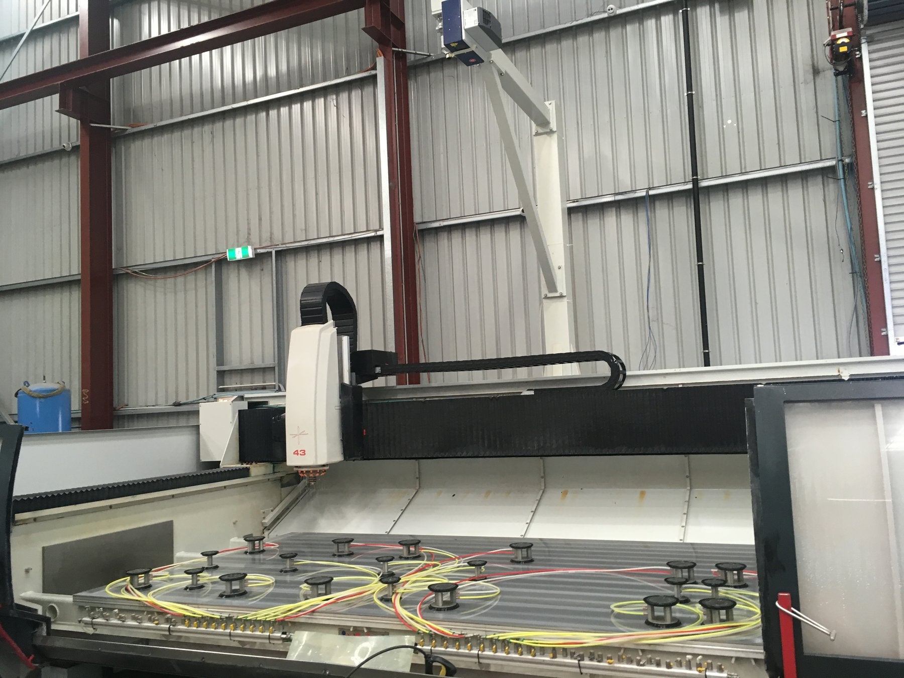 A CNC router bed fitted with vacuum suction pods and stop pins, positioned under an overhead laser projector ahead of a stone or ceramic slab being loaded